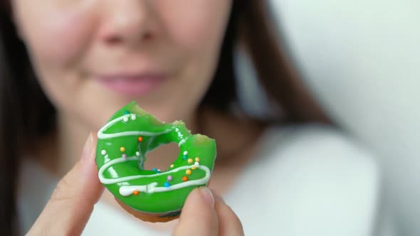 The Girl Brings Her Hand To Her Left Cheek Looking at a Bitten Donut with a Beautiful Pastry