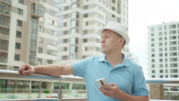 Man in a Hat and with a Phone in His Hand Stands Near a Metal Fence on a City Street