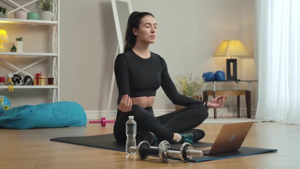 Relaxed Caucasian Sportswoman Sitting on Exercise Mat in Lotus Pose with Laptop at Front. Portrait