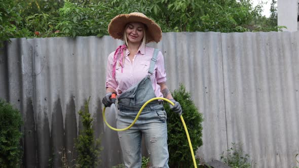 A Young Adult Woman Wateres a Vegetable Garden with a Garden Hose Outside the House