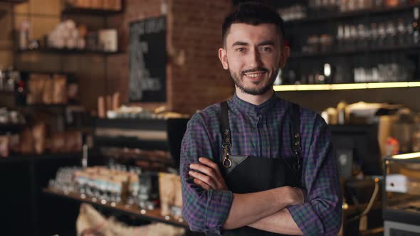 Portrait of Caucasian Barista Guy Wearing Apron Smiling and Standing with Arms Crossed Inside Bar or
