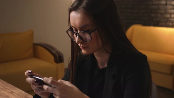 Portrait of Young Woman in Glasses Plays Mobile Game on Phone at Home or Office