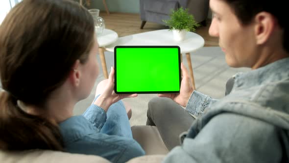 Over The Shoulder View Young Couple at Home Sitting on a Sofa Using with Green Mock-up Screen Tablet