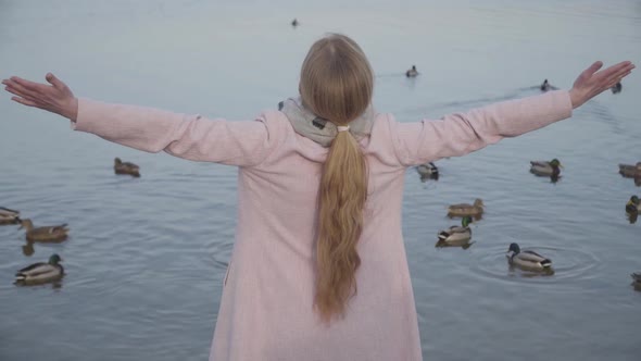 Back View of Adult Caucasian Blond Woman in Pink Coat Stretching Hands at the Background of Calm