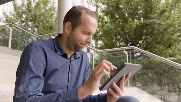 A Caucasian Man Acts Unhappy and Shakes His Head As He Works on a Tablet and Sits on a Staircase