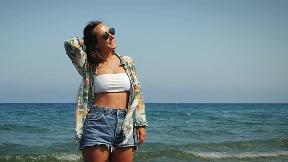 Cheerful carefree woman in summer tropical shirt and jeans shorts standing on seashore
