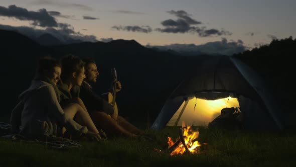 Group of Three Friends Play Guitar and Sing at Camp Fire in Nature Mountain Outdoor Camping Scene at