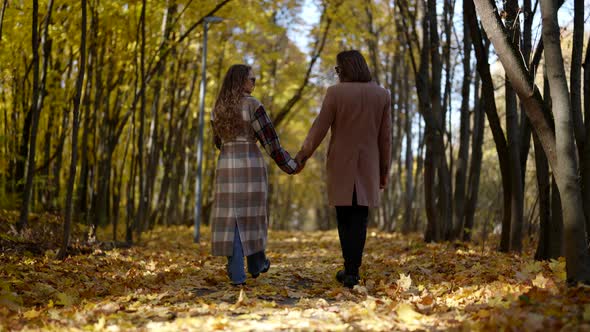 a Man and a Woman in a Coat and Dark Glasses Hold Hands and Walk Along the Fallen Yellow Leaves