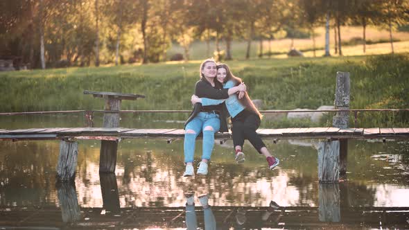 Two Friends are Sitting and Hugging on the Bridge By the River