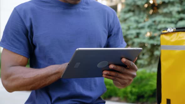 Close Up Afroamerican Man Courier Food Delivery Sitting on Bench Uses Tablet