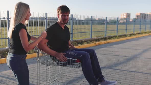 Young Woman Rolls Attractive Guy in the Trolley on Parking Lot of the Supermarket at Sunset