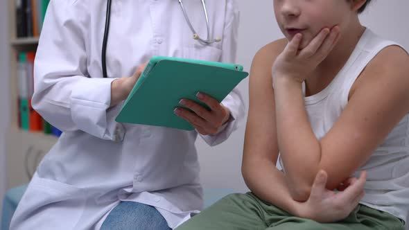 Unrecognizable Caucasian Doctor and Boy Sitting in Hospital with Tablet Talking