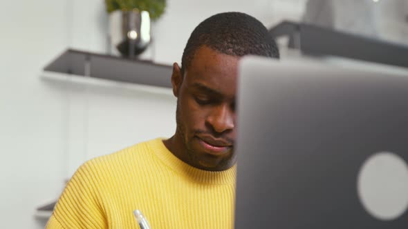 Young african male student taking notes in notebook using laptop at home