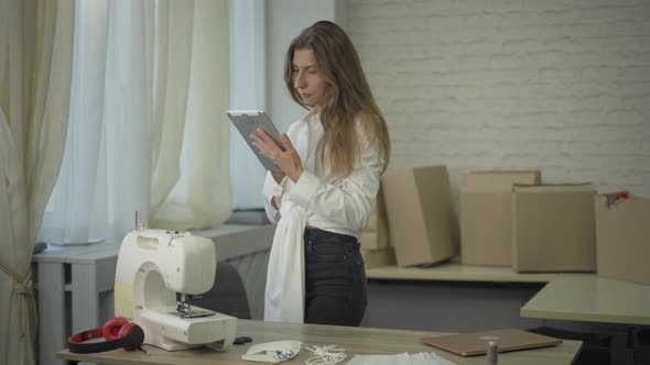 Young Caucasian Woman Entering Room with Tablet and Thinking. Female Seamstress Considering Business