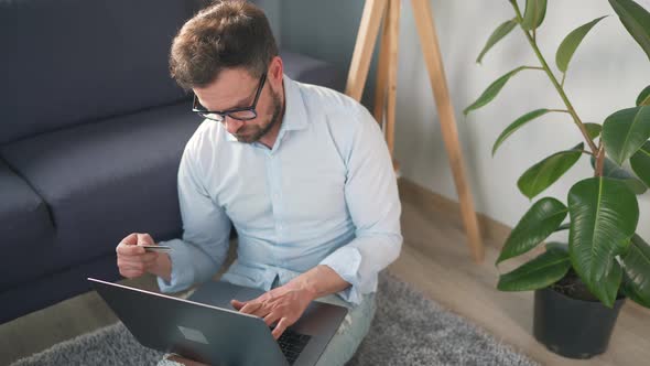 Man with Glasses Sitting on the Floor and Makes an Online Purchase Using a Credit Card and Laptop