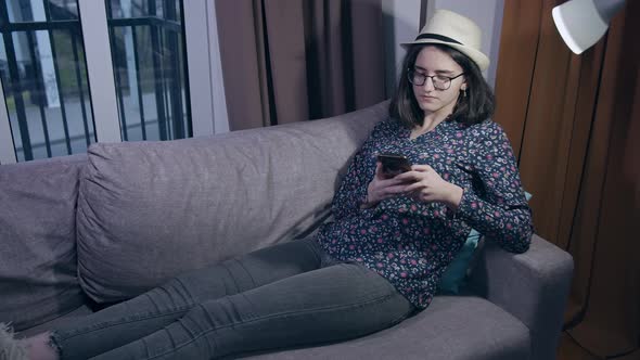 Girl teenager in glasses and a hat reads messages in her mobile phone while sitting on the couch