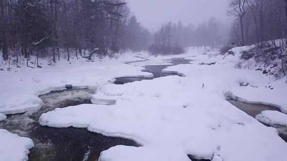 SLOW MOTION aerial shot flying further upstream along an icy river during a winter snow storm