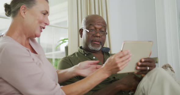 Happy senior diverse couple wearing shirts and using tablet in living room