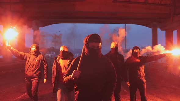 Group of Young Men in Balaclavas with Red Burning Signal Flare Walking on the Road Under the Bridge