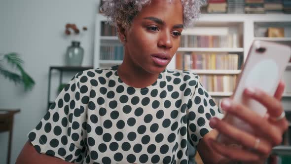 Young African American Woman Looks at Phone Sits at Table in Living Room