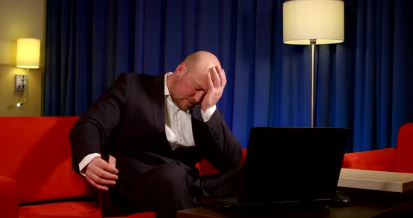 a Bald Man in a Dark Suit Is Sitting on a Red Sofa in Front of a Laptop Against a Blue Curtain. He