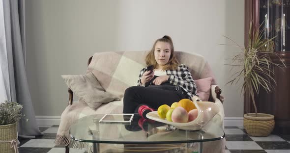 Portrait of Bored Caucasian Teenage Girl Sitting on Couch and Switching Channels