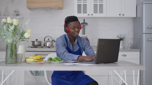 Young Positive Man in Earphones and Apron Sitting at Kitchen Table Typing on Laptop Keyboard