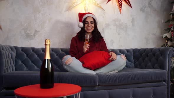 Young Woman in Santa Claus Hat with Glass of Champagne Sitting on Sofa in Room