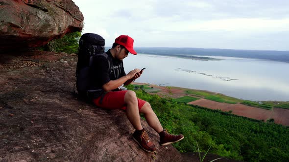 hiker man sitting and using smartphone on the edge of cliff, on a top of the rock mountain