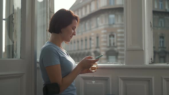 Side View of Young Woman Uses a Smartphone While Drinking Wine and Sitting By the Window at Home on