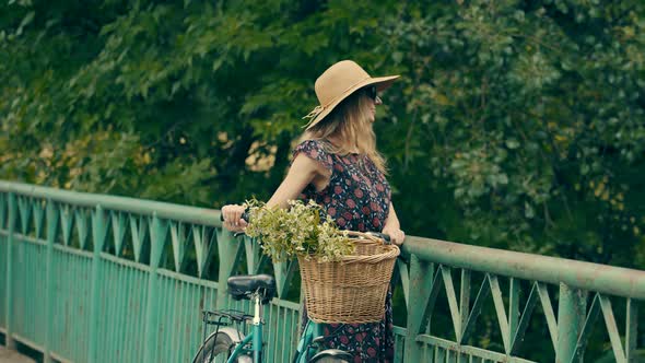 Girl Walking With Bicycle On The Bridge. Woman In Hat. Wicker Basket With Flowers.