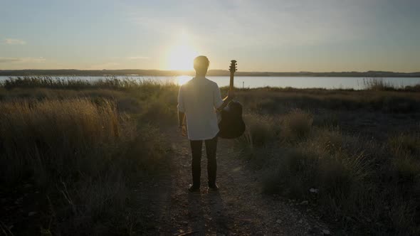 Silhouetted Man with Guitar Jumping and Clapping His Legs Against a Sunset Sun  Slowmotion Back View