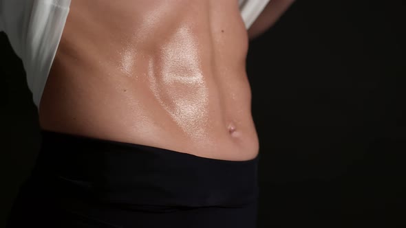 Woman Lifts a White Tshirt with Hands and Shows Muscular Sweaty Belly Closeup Against a Darkness