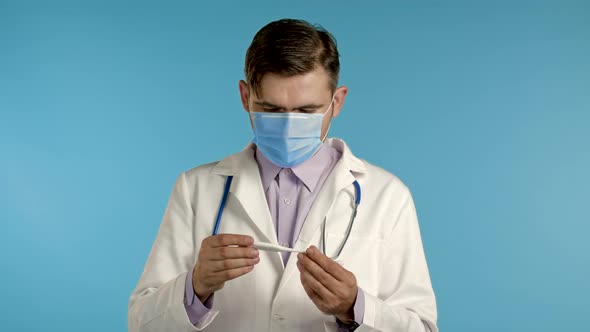 Doctor in Medical Coat and Mask Looks at Thermometer and Shows with Gesture That He Doesn't Know