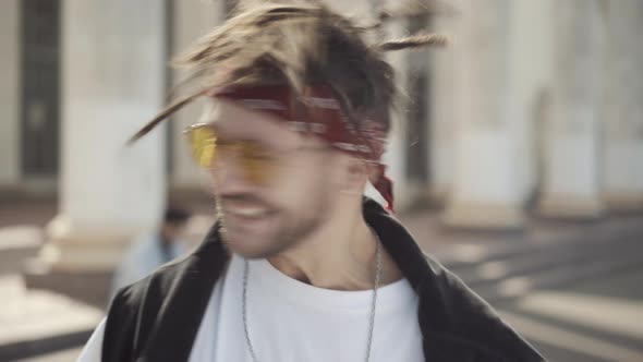 Headshot of Joyful Caucasian Hippie in Sunglasses Shaking Dreadlocks and Smiling at Camera