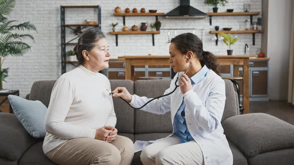 Woman Doctor Examining Aged Lady By Stethoscope While She is Breathing Deeply Sitting on Couch