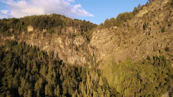 Aerial dolly in flying over pine tree woods and Corbata Blanca waterfall between mountains at sunset