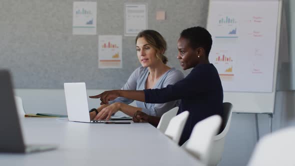 Two diverse female colleagues sitting at desk with laptop and discussing in office