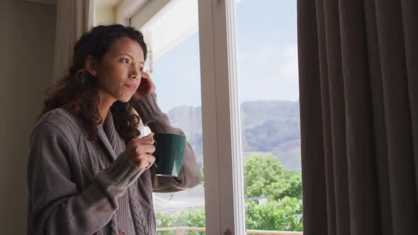 Thoughtful biracial woman standing with coffee at window and looking into distance