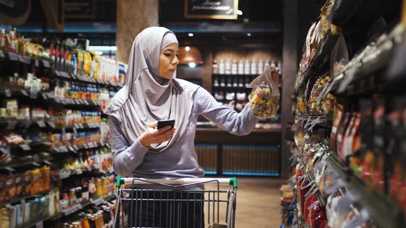 Portrait of Young Beautiful Muslim Woman in Hijab Using Phone and Choosing Food in the Supermarket