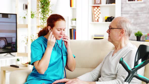 Caucasian Female Nurse Listening To Elderly Retired Man Heartbeat in Bright and Cozy Nursing Home alt