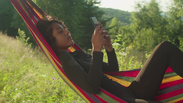 Pretty Relaxed African American Woman Lying in Hammock Networking Online on Phone in Mountains