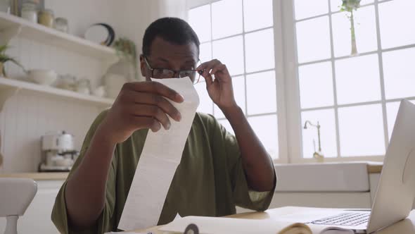 African American Man Looks at Checks Near Laptop in Kitchen