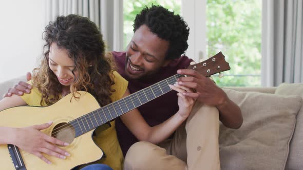 Happy biracial couple sitting on sofa together and playing guitar