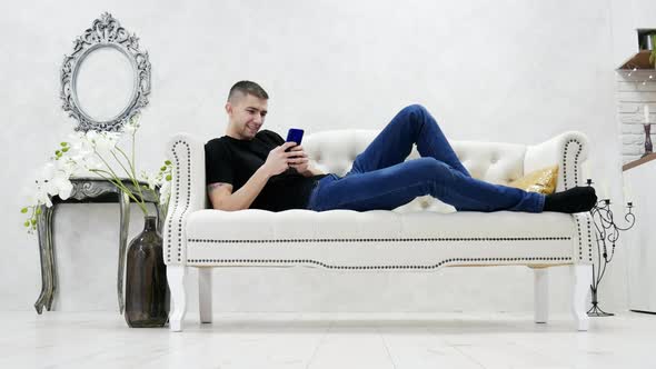 Young Man Relaxing on Couch and Using Smartphone in Modern Luxury House, Chatting with Friends