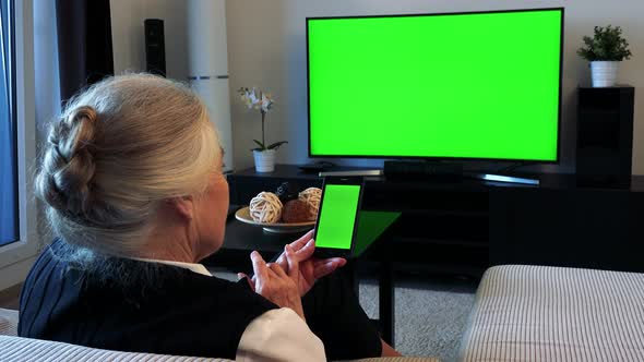 Old Caucasian Woman Watches Television in Living Room and Works on Smartphone