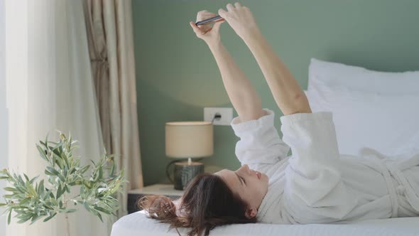 Pretty smiling brunette woman in white bathrobe making selfie by phone and lying on bed at home