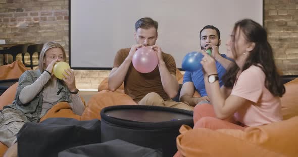 Relaxed Multiethnic Young Friends Sitting on Bag Chairs and Blowing Balloons. Portrait of Cheerful