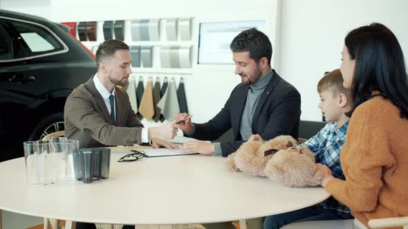 Businessman Buying Car in Dealership Signing Sales Contract Indoors with Wife and Son