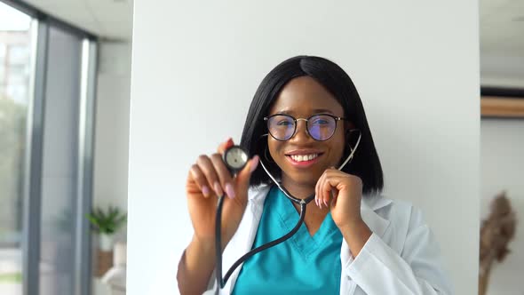 African American Woman Doctor in White Coat in Clinic at Workplace Holds Stethoscope in His Hand and